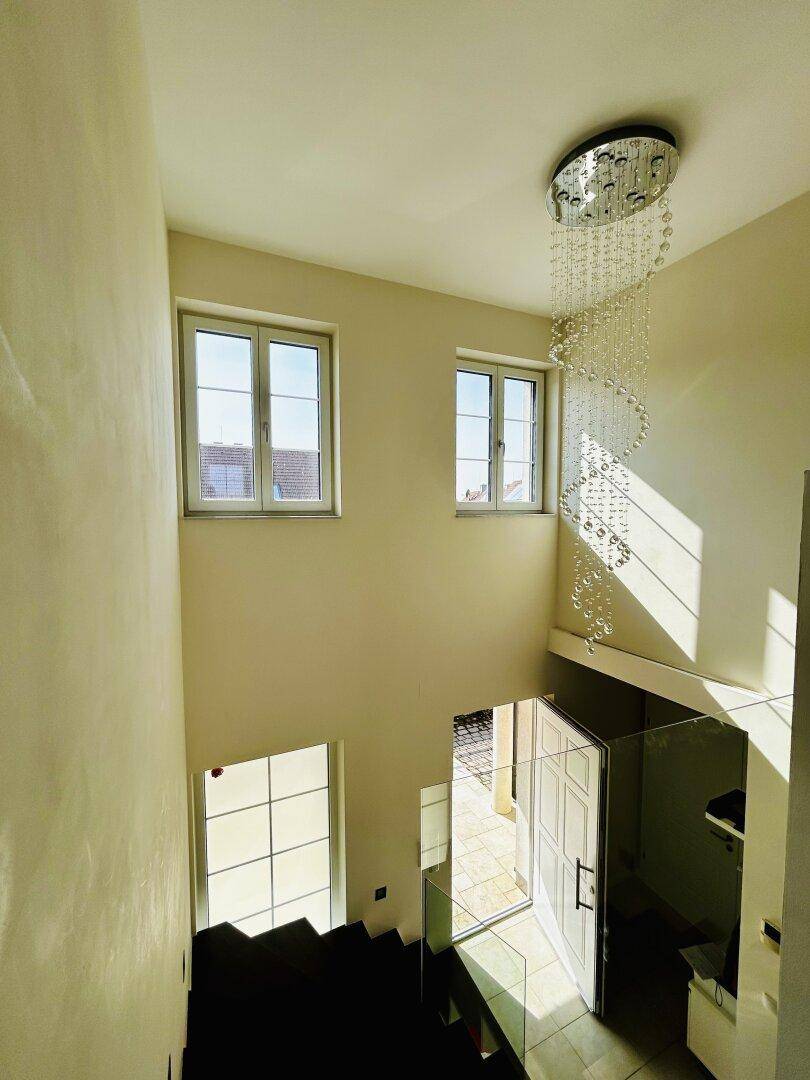 Treppenhaus mit moderner Hängeleuchte, großen Fenstern und viel Tageslicht.