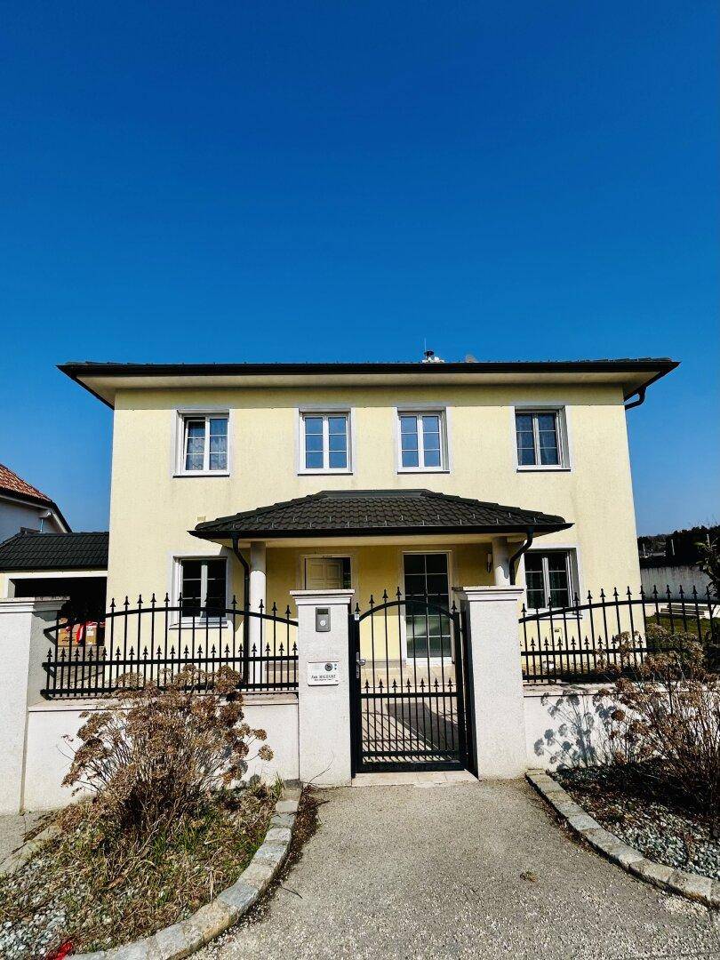 Vorderansicht eines gepflegten Einfamilienhauses mit gelber Fassade und schmiedeeisernem Zaun.