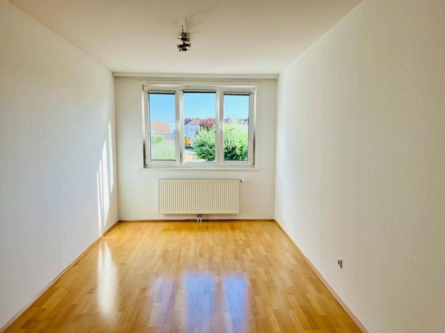 Leeres Zimmer mit hellem Parkettboden und großem Fenster für viel Tageslicht.