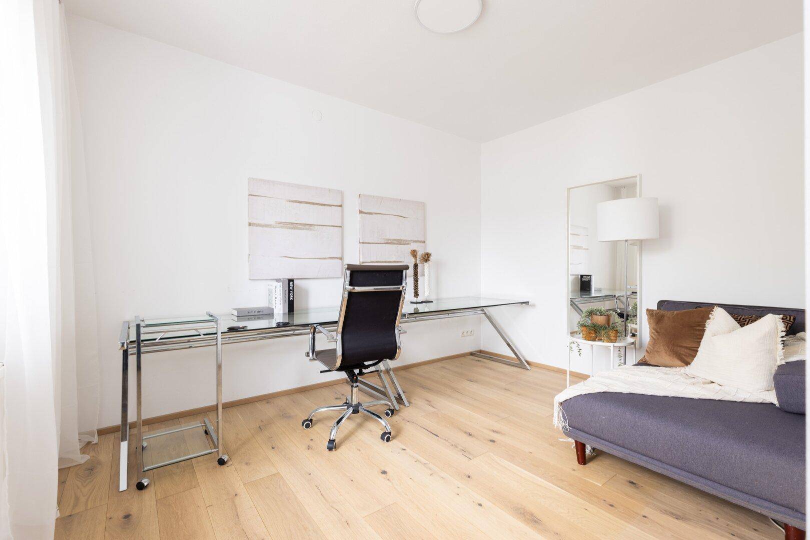 Helles Arbeitszimmer mit modernem Glastisch, Bürostuhl und bequemer Liege.