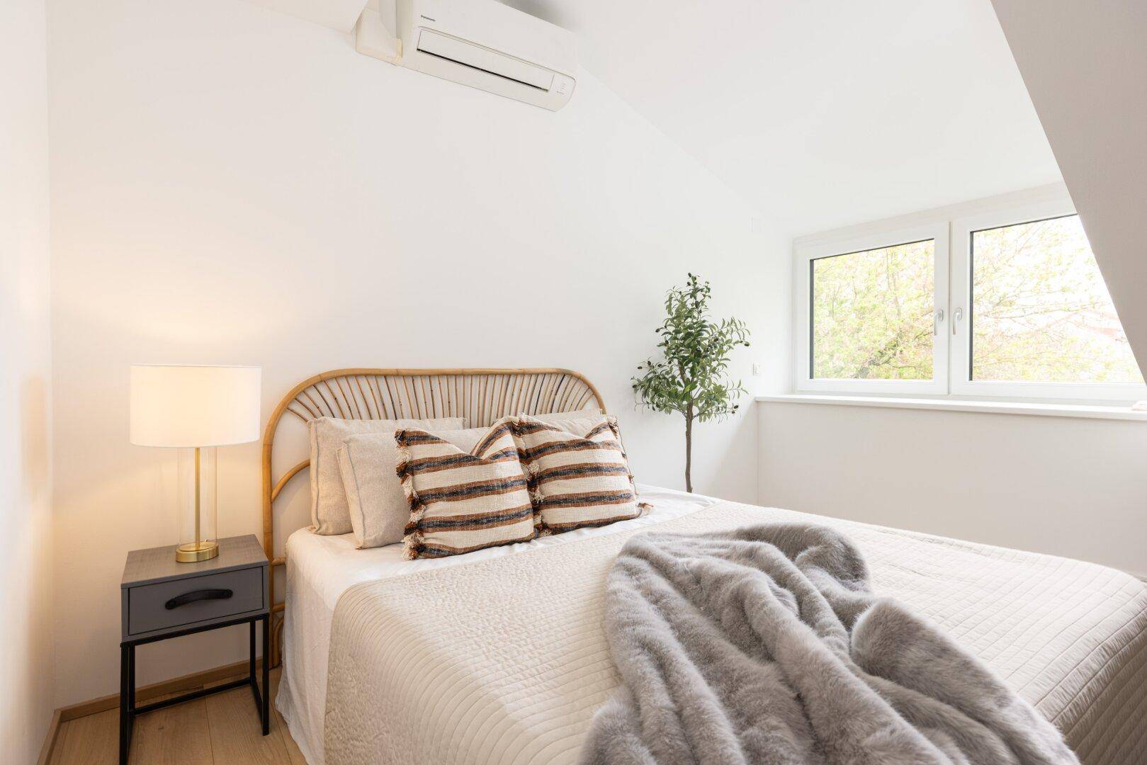 Schlafzimmer im Dachgeschoss mit Bett, Nachttisch, Fenster und Klimaanlage.