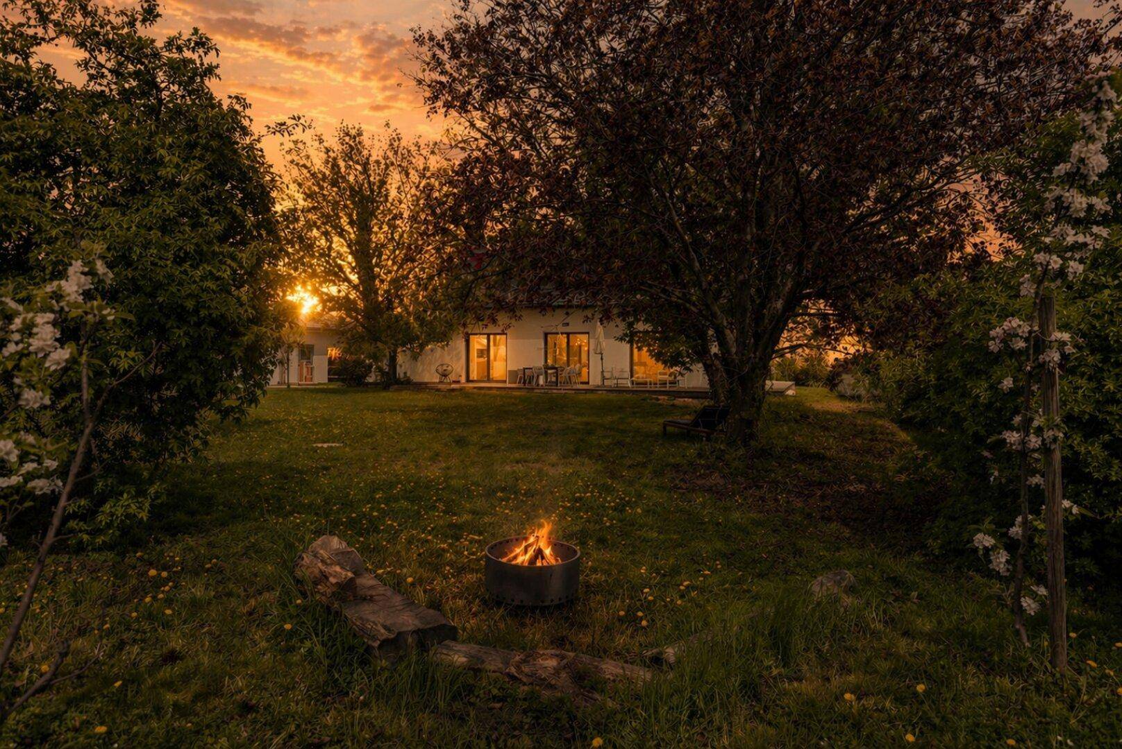 Abendliche Stimmung im Garten mit Feuerstelle, Bäumen und beleuchtetem Haus.