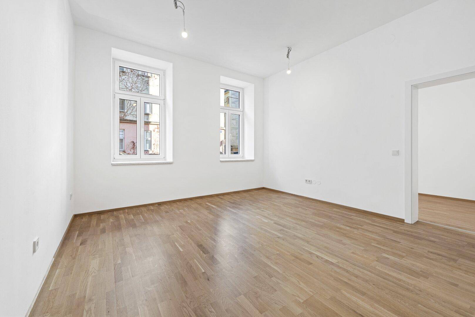 Leerer Raum mit hellem Holzboden und zwei großen Fenstern, bereit zur Einrichtung.