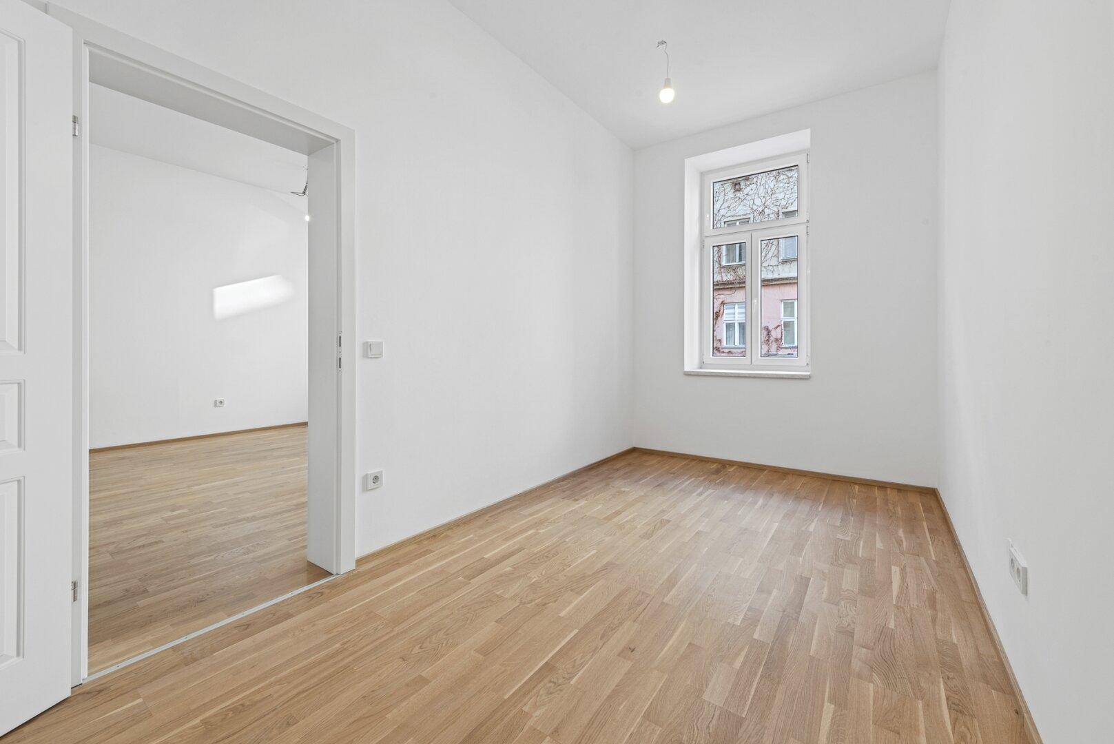 Leeres Zimmer mit Holzboden und einem großen Fenster, das viel Tageslicht hereinlässt.