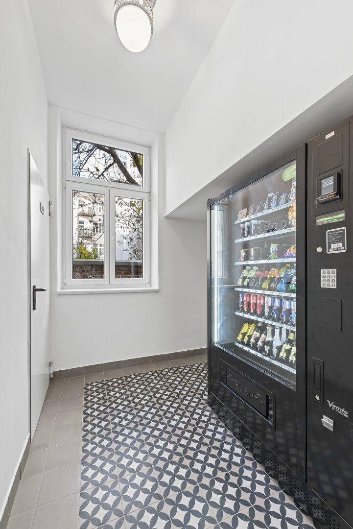 Heller Flur mit Fenster, gemusterten Bodenfliesen und einem Verkaufsautomaten.