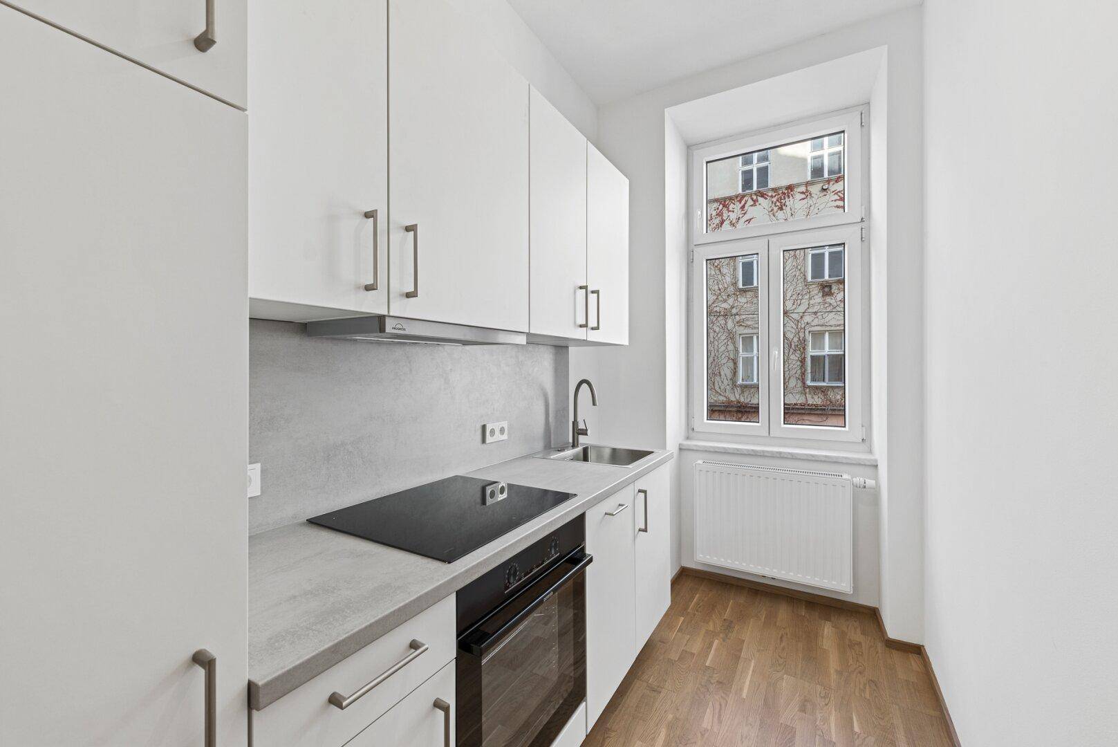 Moderne Küchenzeile mit weißen Schränken, integriertem Kochfeld und Backofen unter dem Fenster.