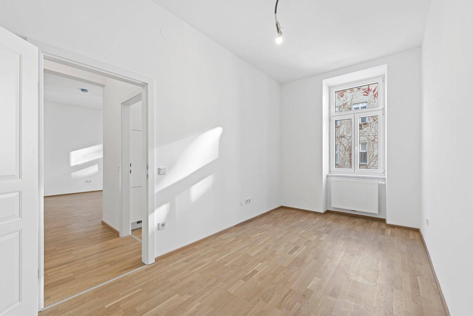 Lichtdurchfluteter Wohnraum mit Parkettboden und Zugang zu einem angrenzenden Zimmer.