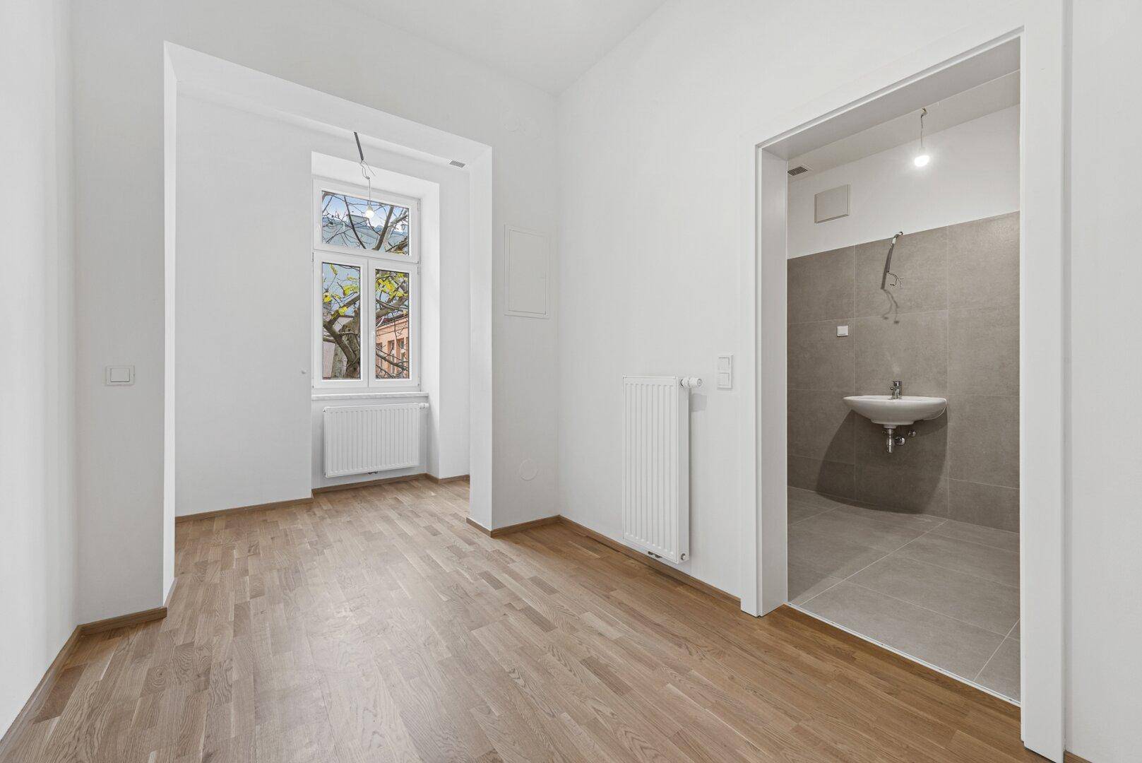 Heller Wohnraum mit Holzparkettboden und Zugang zu einem Badezimmer und einem weiteren Zimmer.