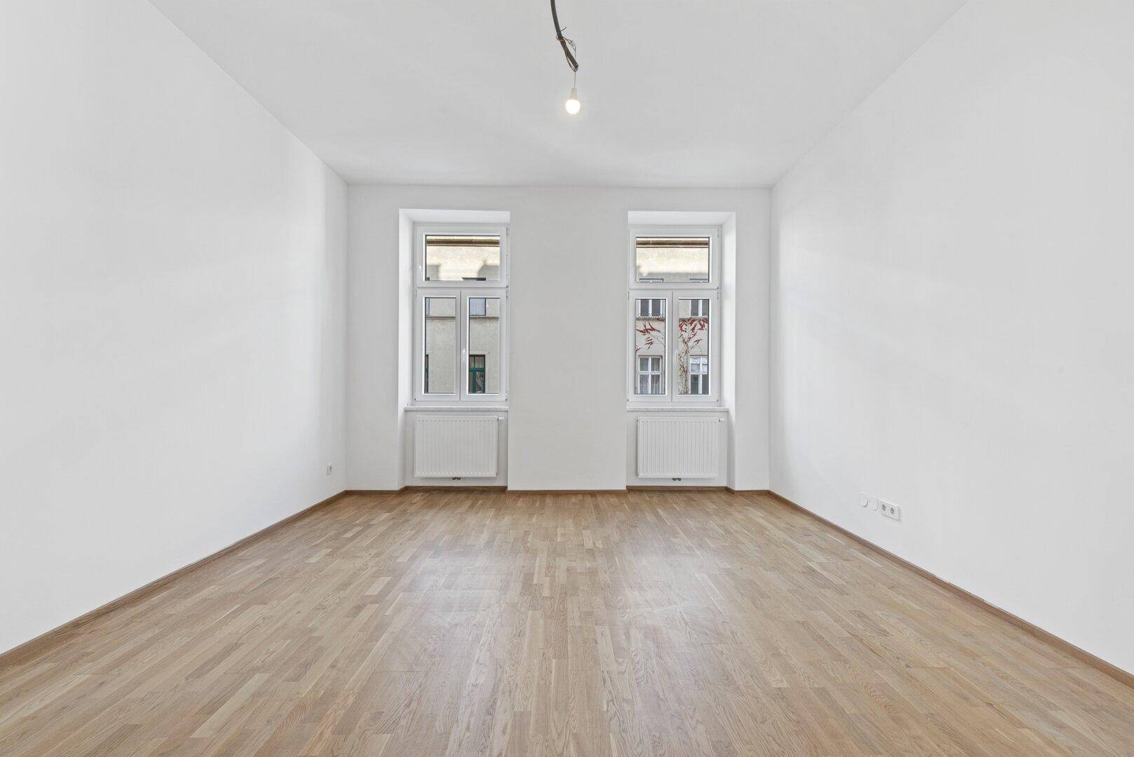 Helles, leeres Zimmer mit Parkettboden und zwei Fenstern, bereit zur individuellen Gestaltung.