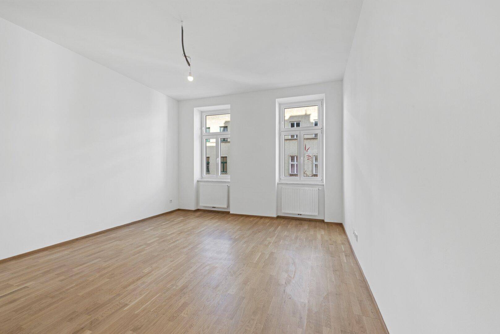 Geräumiges, leeres Zimmer mit hellem Parkettboden und zwei großen Fenstern.