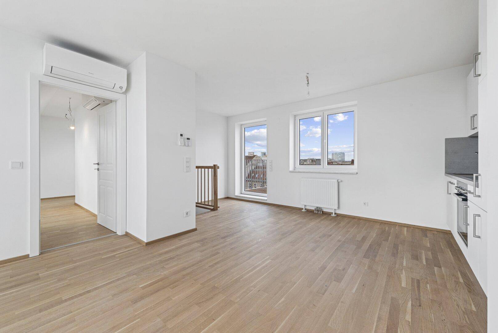 Heller offener Wohnbereich mit Holzboden, Zugang zu einem Balkon und integrierter Küche, mit Klimaanlage.