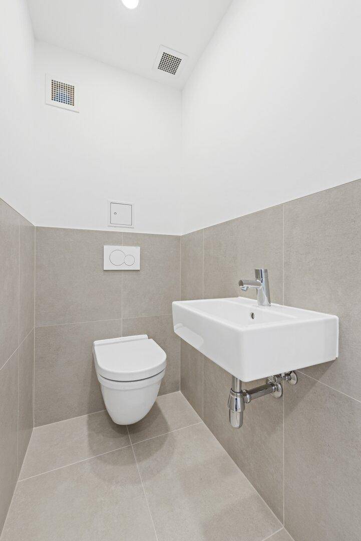 Modernes Gäste-WC mit grauen Fliesen, wandhängendem WC und kleinem Waschbecken.