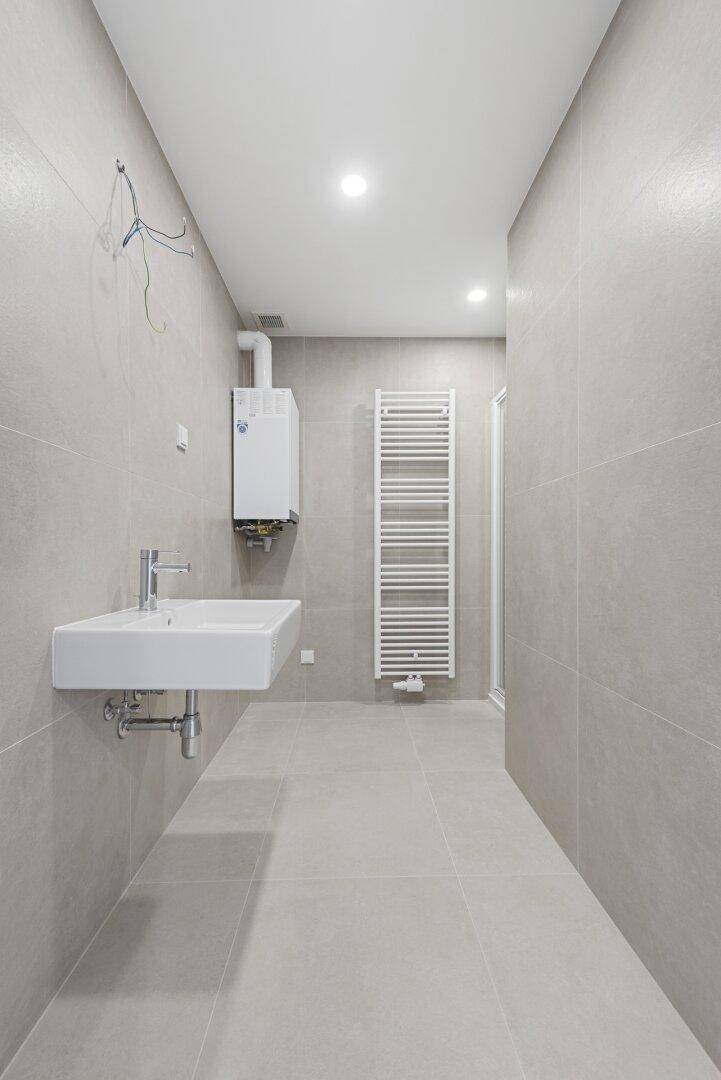 Funktionales Badezimmer mit grauen Fliesen, Waschbecken, beheiztem Handtuchhalter und Boiler.