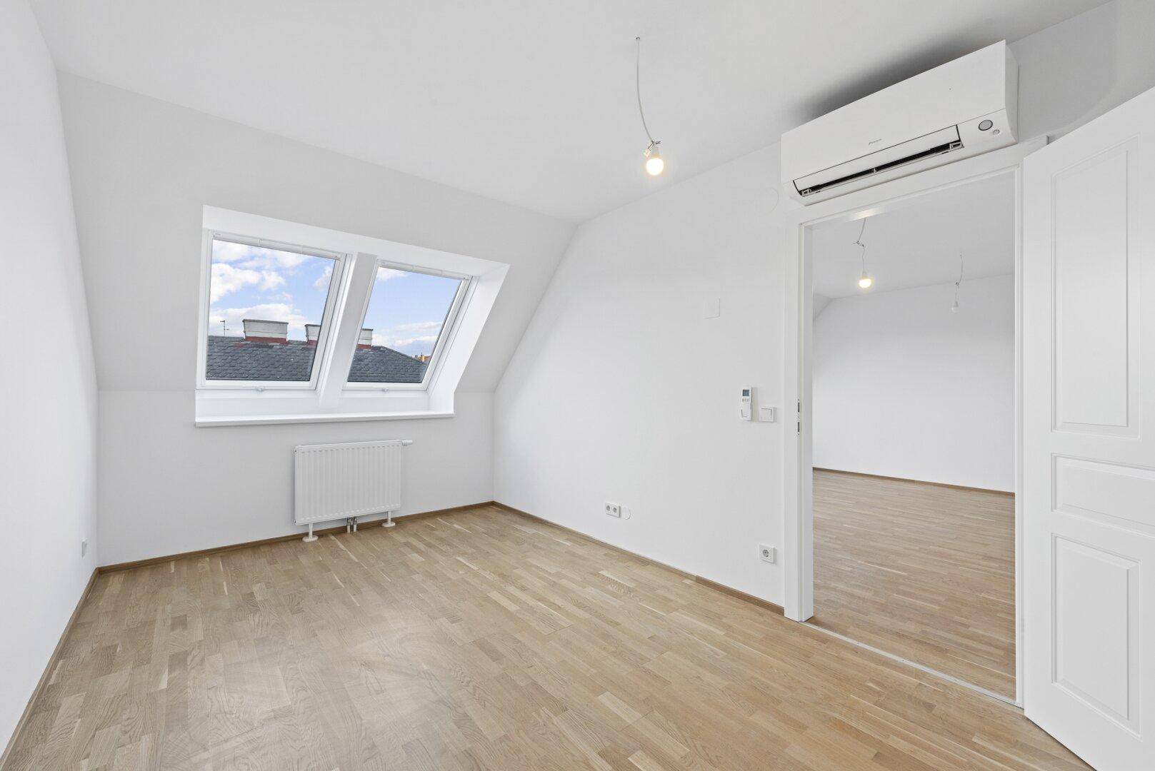 Helles Schlafzimmer im Dachgeschoss mit Holzparkett, zwei Dachfenstern und Klimaanlage.