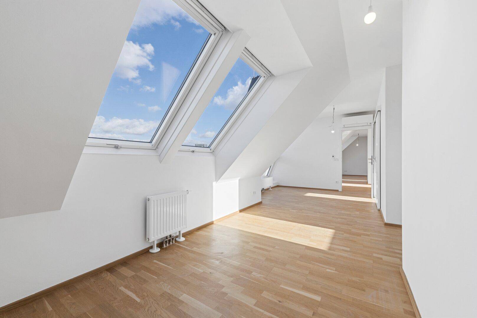 Heller Dachgeschossraum mit schrägen Wänden, Holzboden und großen Dachfenstern mit Himmelsblick.