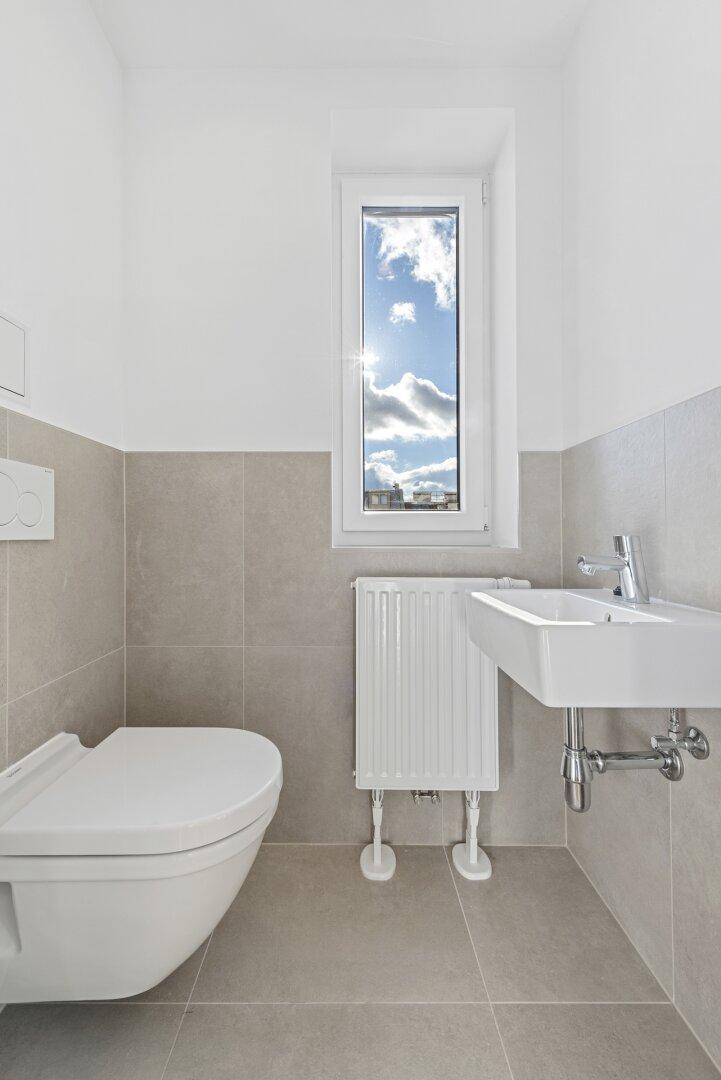 Modernes Gäste-WC mit hellgrauen Fliesen, wandhängendem WC und Fenster.