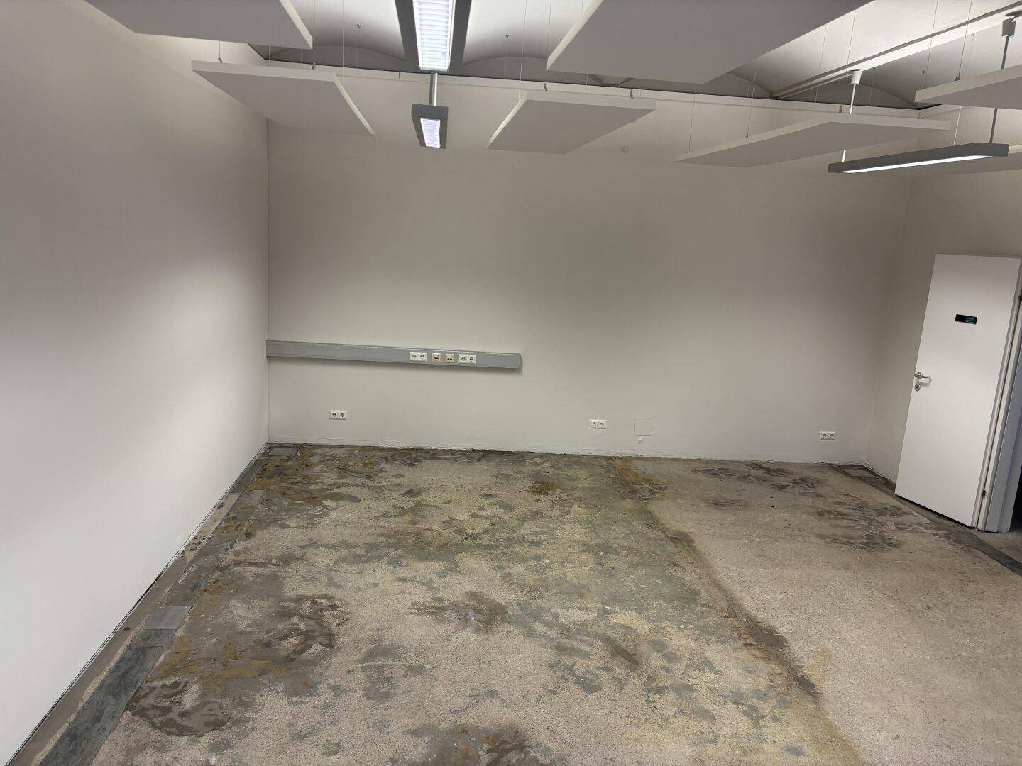 Leerer Büroraum mit Betonboden, schallabsorbierenden Deckenplatten und praktischer Wandsteckdosenleiste.