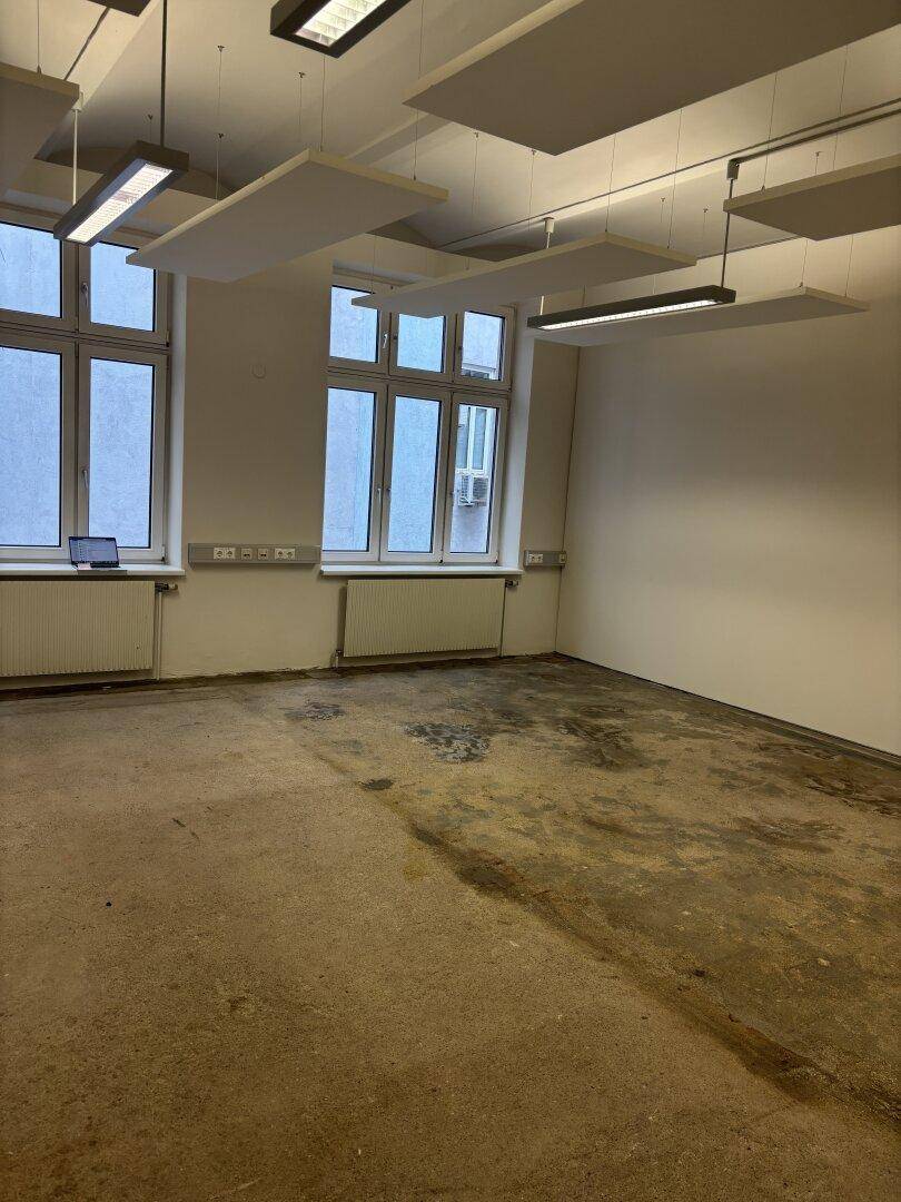 Geräumiger Büroraum mit großen Fenstern, Betonboden und schallabsorbierenden Deckenplatten, bietet viel Tageslicht.