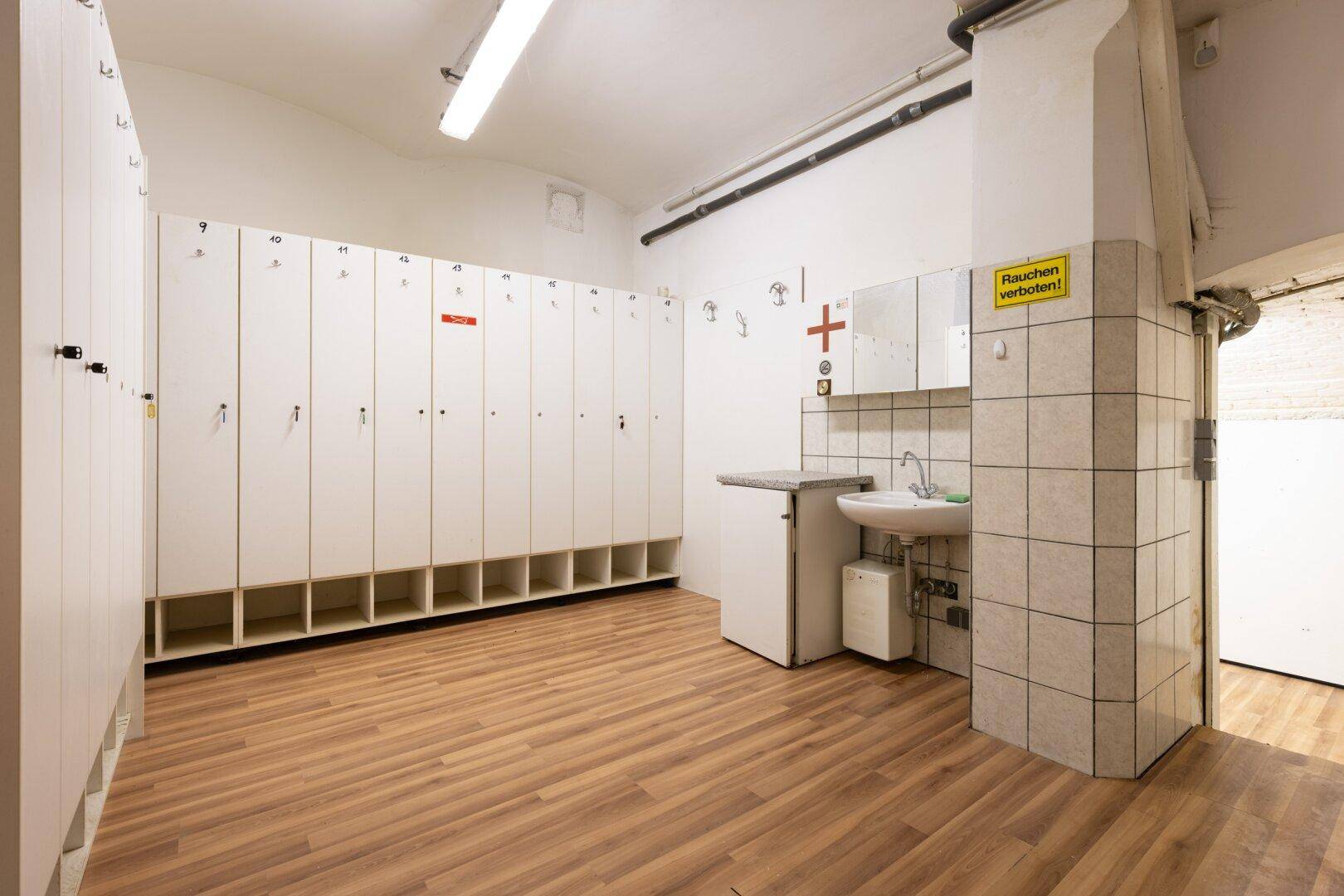 Umkleideraum mit vielen Spinden, Waschbecken und gefliesten Wänden, mit Holzoptikboden.