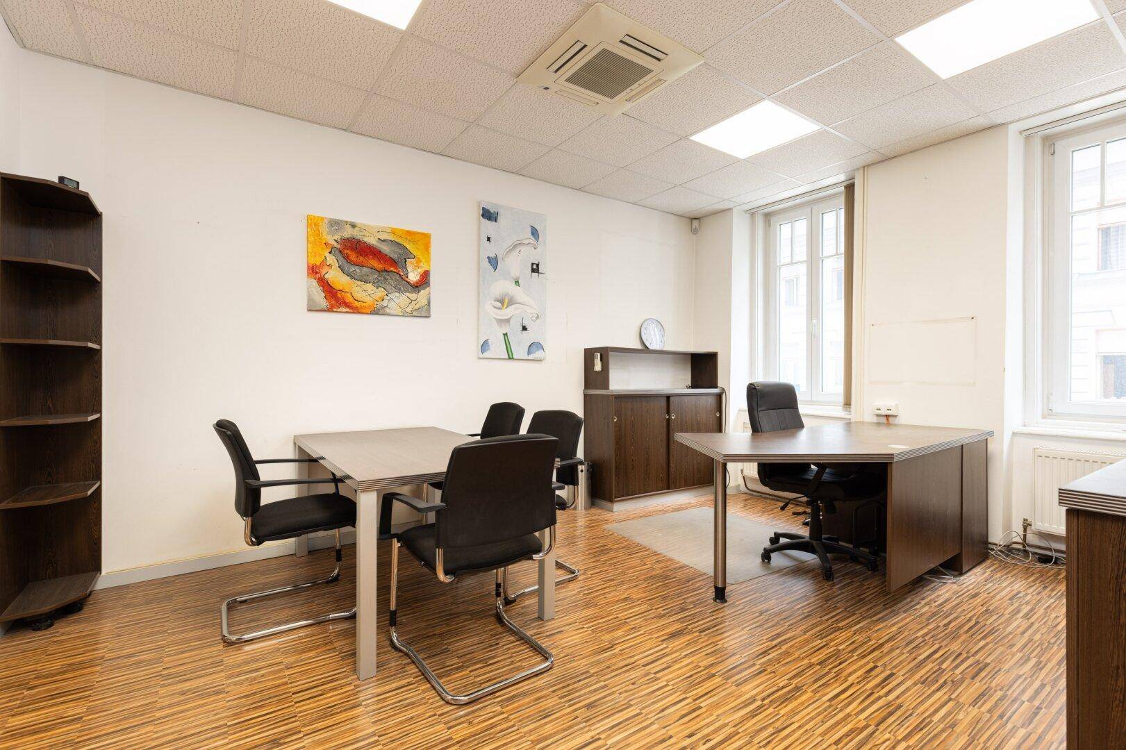 Geräumiges Büro mit mehreren Schreibtischen, Holzoptikboden und farbenfrohen Bildern an der Wand.