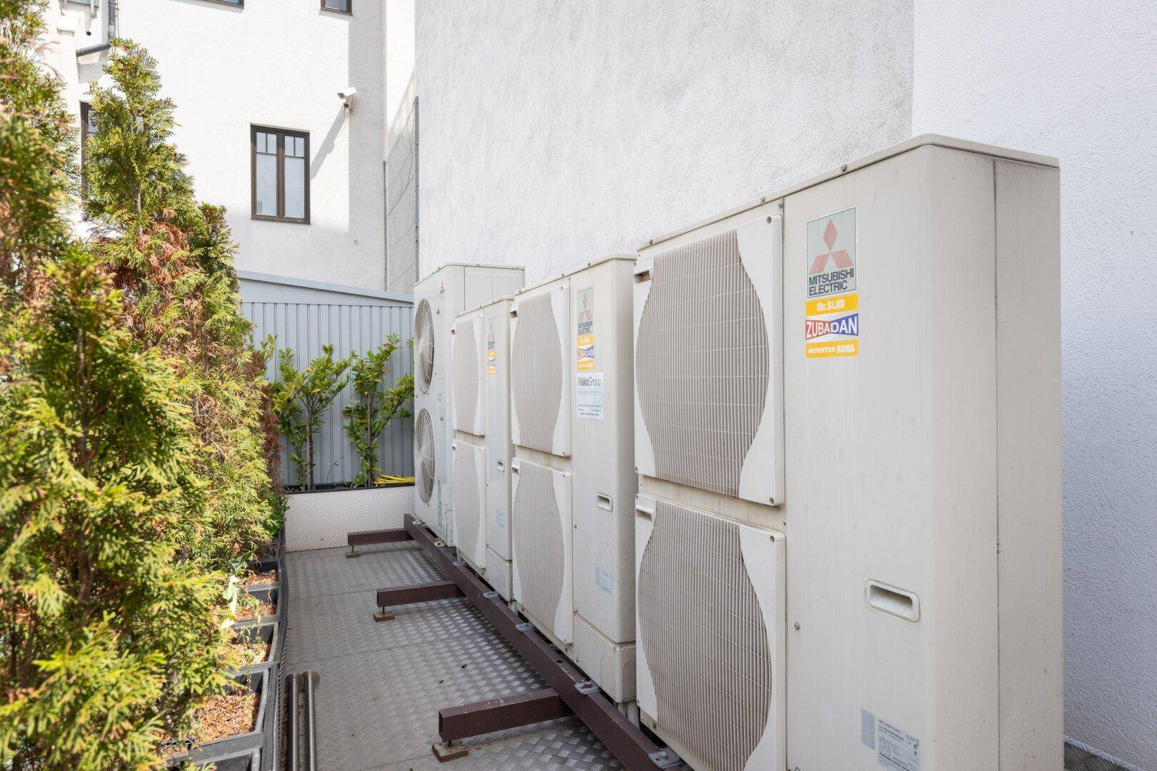 Reihe von Klimaanlagen-Außengeräten an der Gebäudewand, umgeben von Pflanzen.
