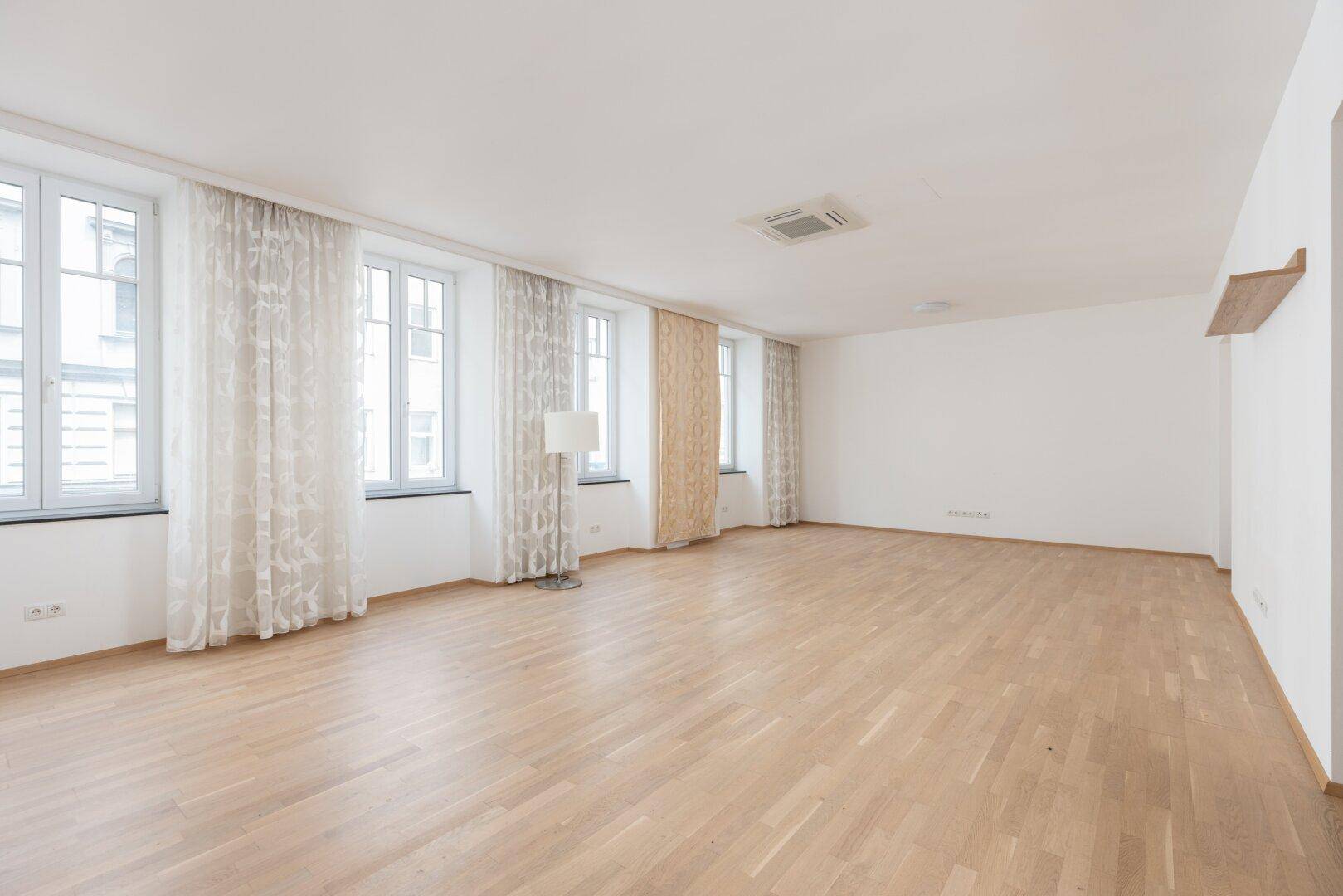 Helles, leeres Wohnzimmer mit Parkettboden, hohen Decken und großen Fenstern.