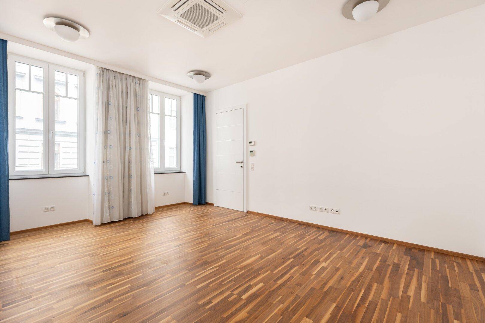 Geräumiges Wohnzimmer mit Parkettboden, weißen Wänden und blauen Vorhängen.
