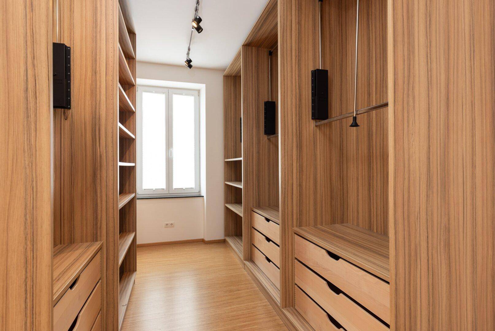 Begehbarer Kleiderschrank mit Holzregalen, Schubladen und Fenster für Tageslicht.