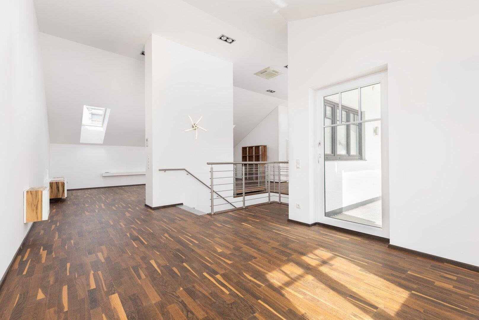 Heller Dachgeschoss-Wohnbereich mit Holzboden, Treppe und Zugang zu einem Balkon.
