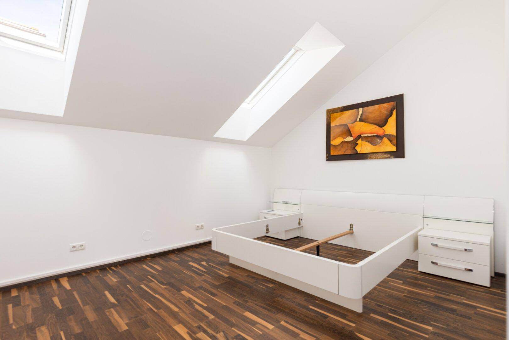 Schlafzimmer im Dachgeschoss mit Holzboden, Dachfenstern und weißem Bettrahmen.