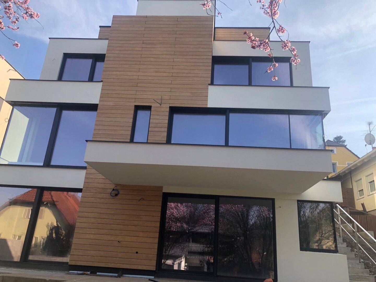 Moderne Gebäudefassade mit großen Fenstern und Holzverkleidung, umgeben von blühenden Bäumen im Frühling.