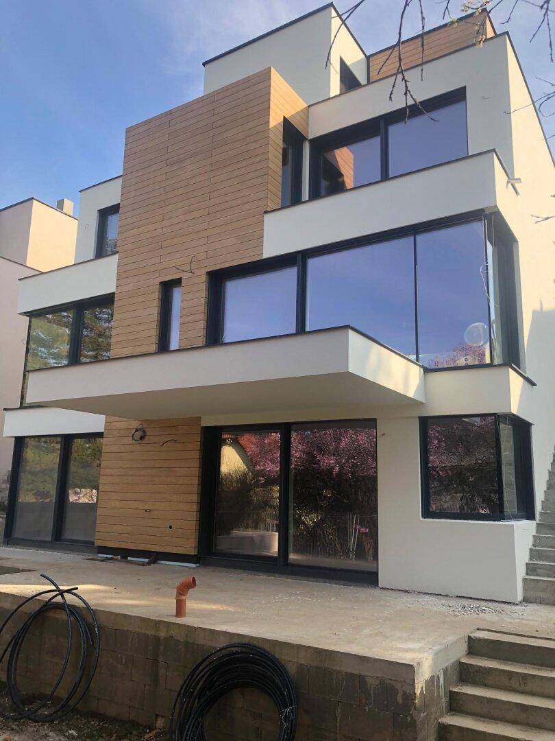 Moderne Gebäudefassade mit großen Glasflächen und Holzdetails, umgeben von Baustellenmaterialien bei Tageslicht.
