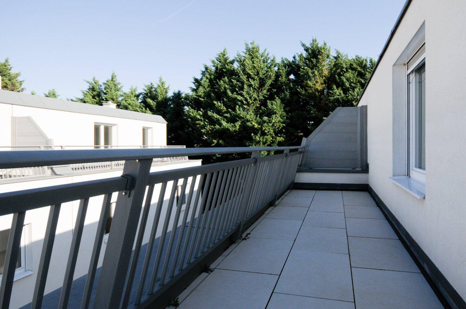 Geräumige Terrasse mit grauen Fliesen und Metallgeländer, umgeben von grünen Bäumen.