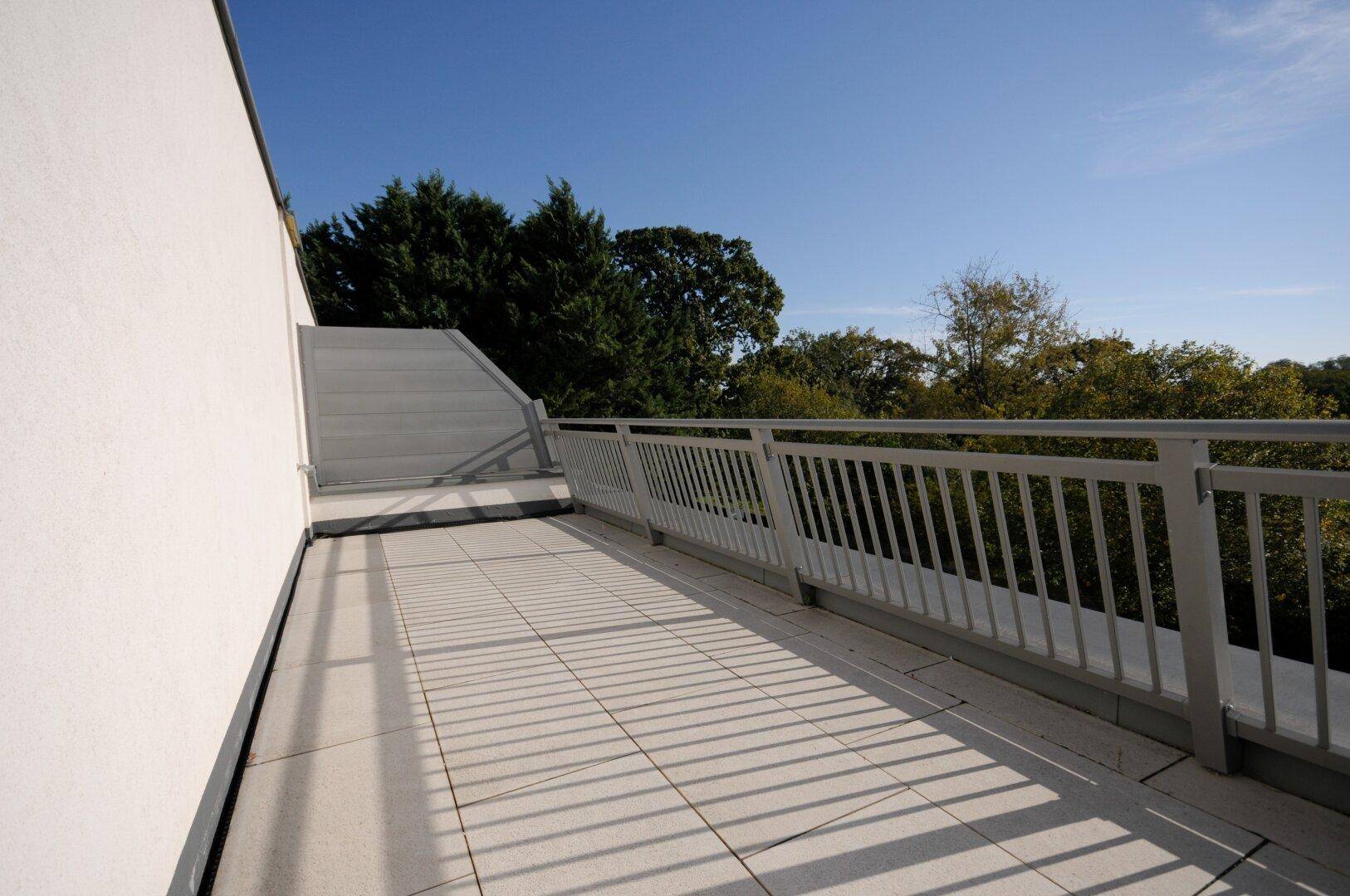 Sonniger Terrassenbereich mit Fliesen und Metallgeländer, umgeben von üppigem Grün und Bäumen.