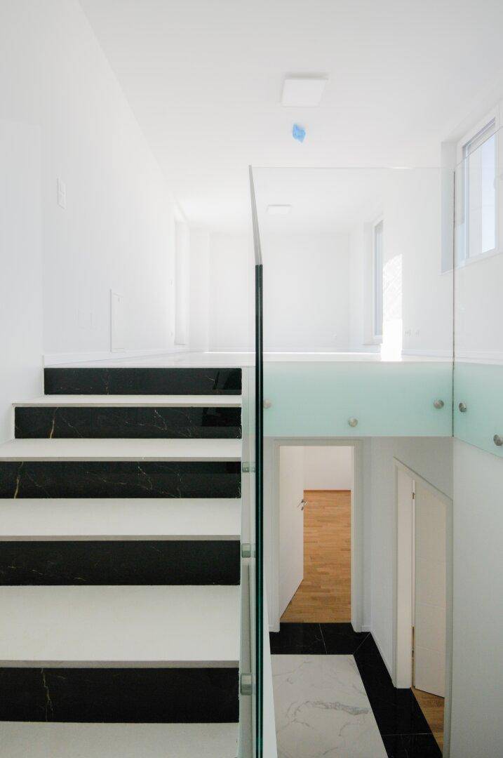Modernes Treppenhaus mit schwarz-weißem Marmor und eleganter Glasbrüstung, führt zu einem hellen Flur.