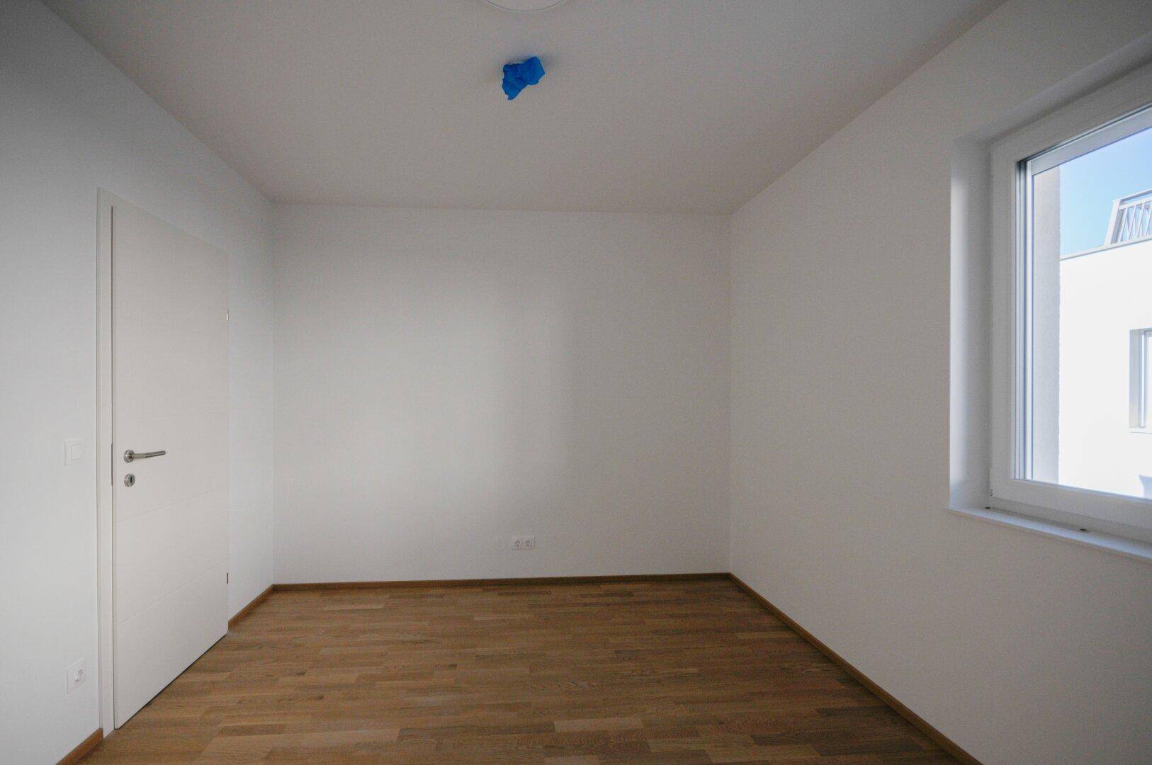 Leeres Zimmer mit hellem Parkettboden, weißen Wänden und einem Fenster mit Blick nach draußen.