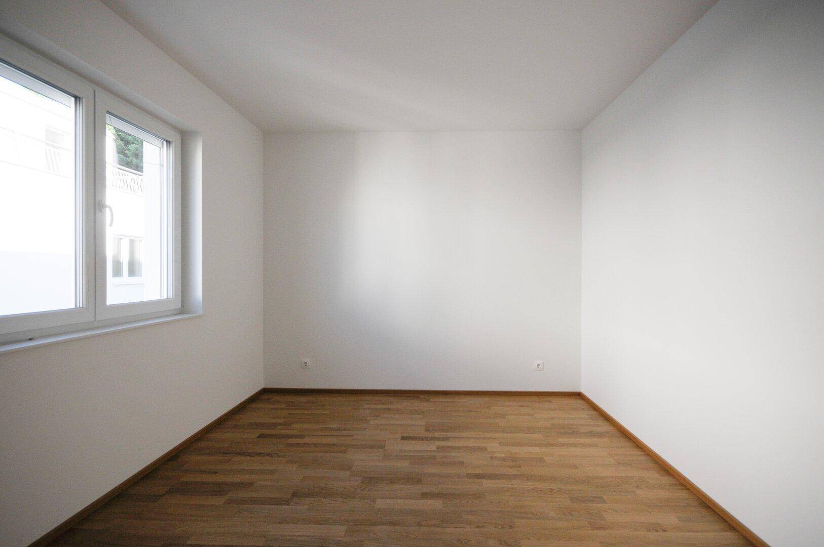 Unmöbliertes Zimmer mit hellem Holzboden, weißen Wänden und großem Fenster für viel Licht.