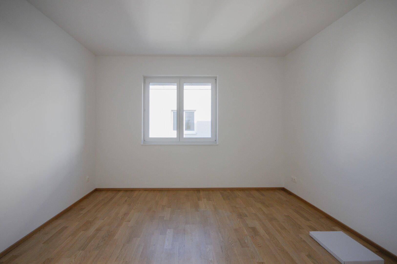 Helles Schlafzimmer mit Parkettboden, weißen Wänden und einem großen Fenster.