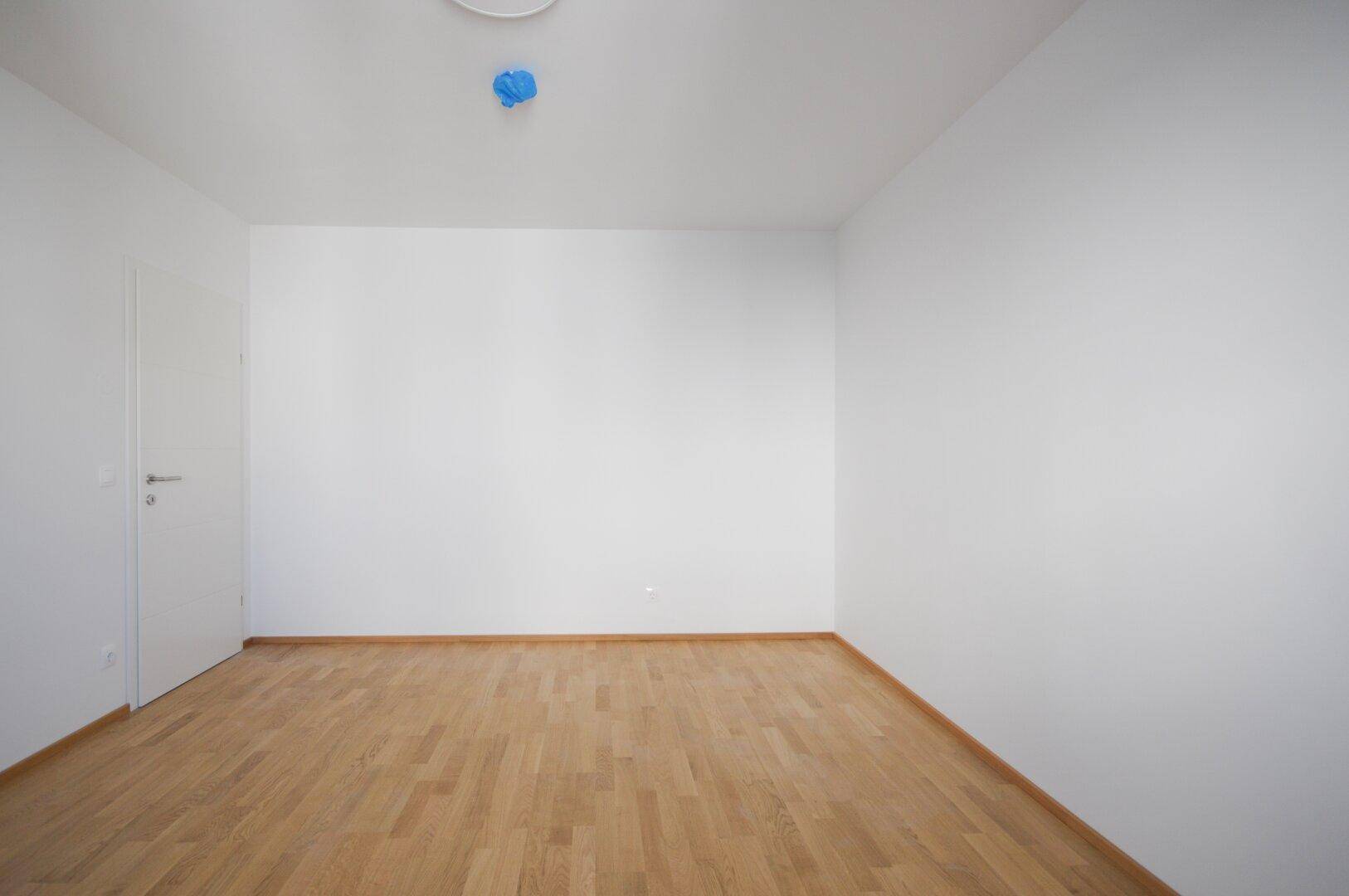 Leeres Zimmer mit hellem Parkettboden, weißen Wänden und einer Tür.