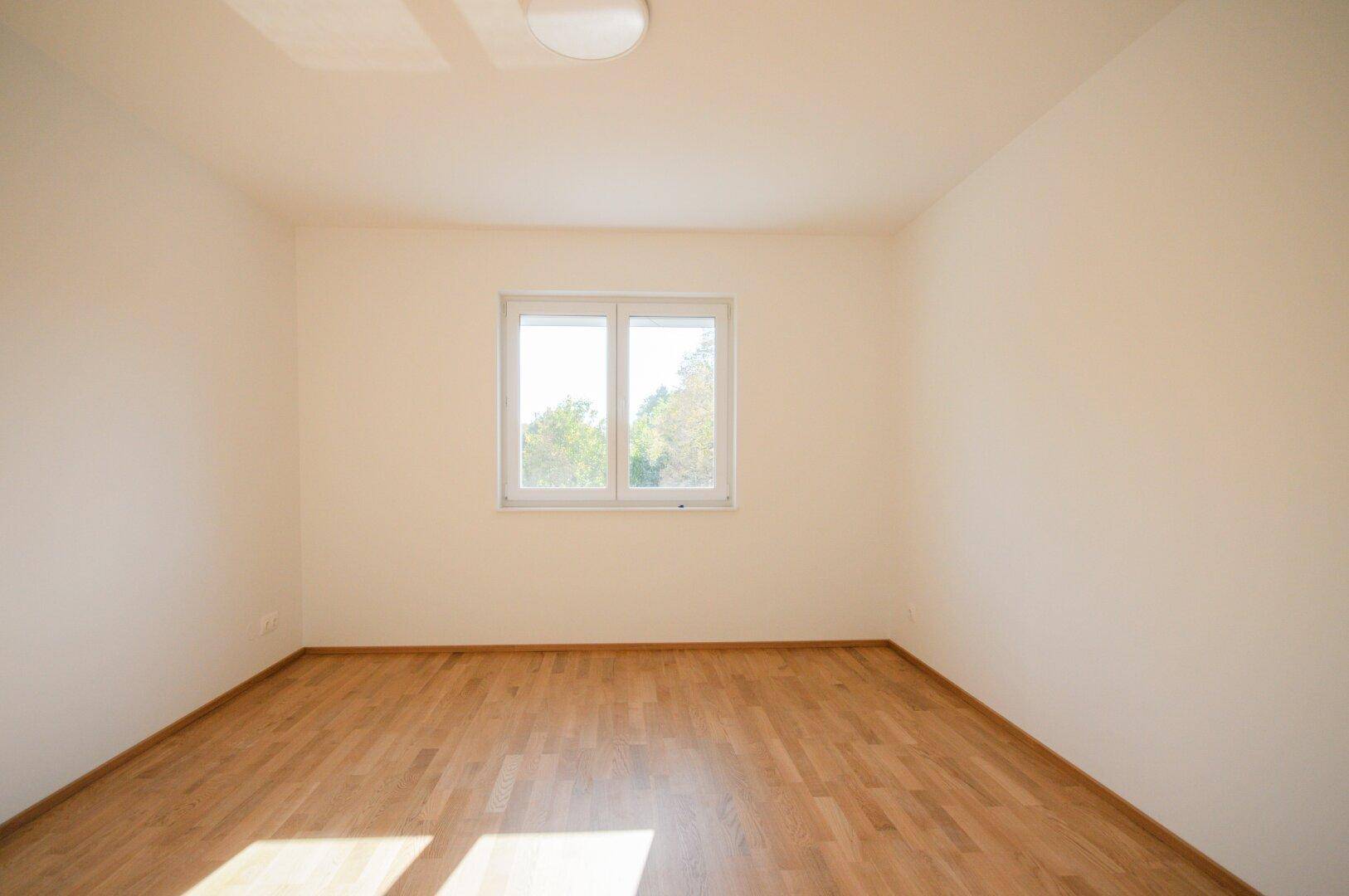 Helles Zimmer mit Parkettboden, weißen Wänden und einem Fenster mit Blick ins Grüne.
