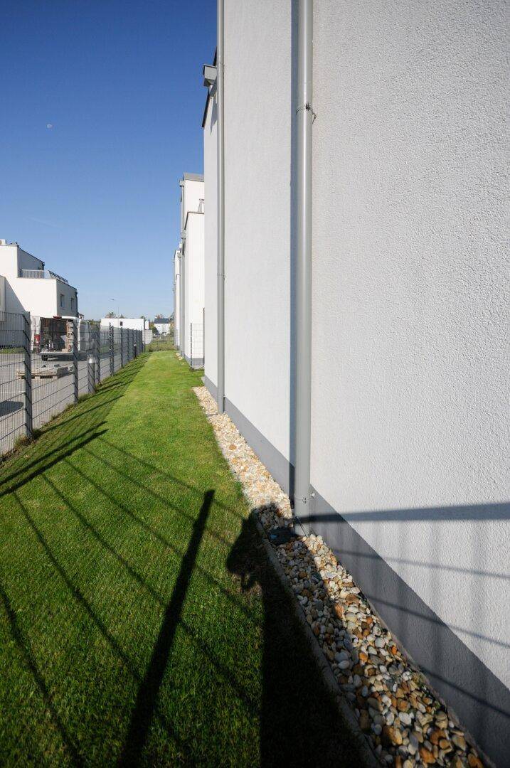 Schmaler Rasenstreifen entlang der Hauswand mit dekorativen Steinen und Metallzaun.