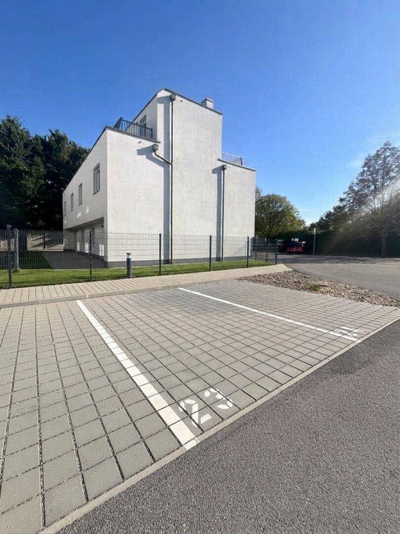 Modernes weißes Gebäude mit Flachdach und gepflastertem Parkplatz, umgeben von einem Zaun.