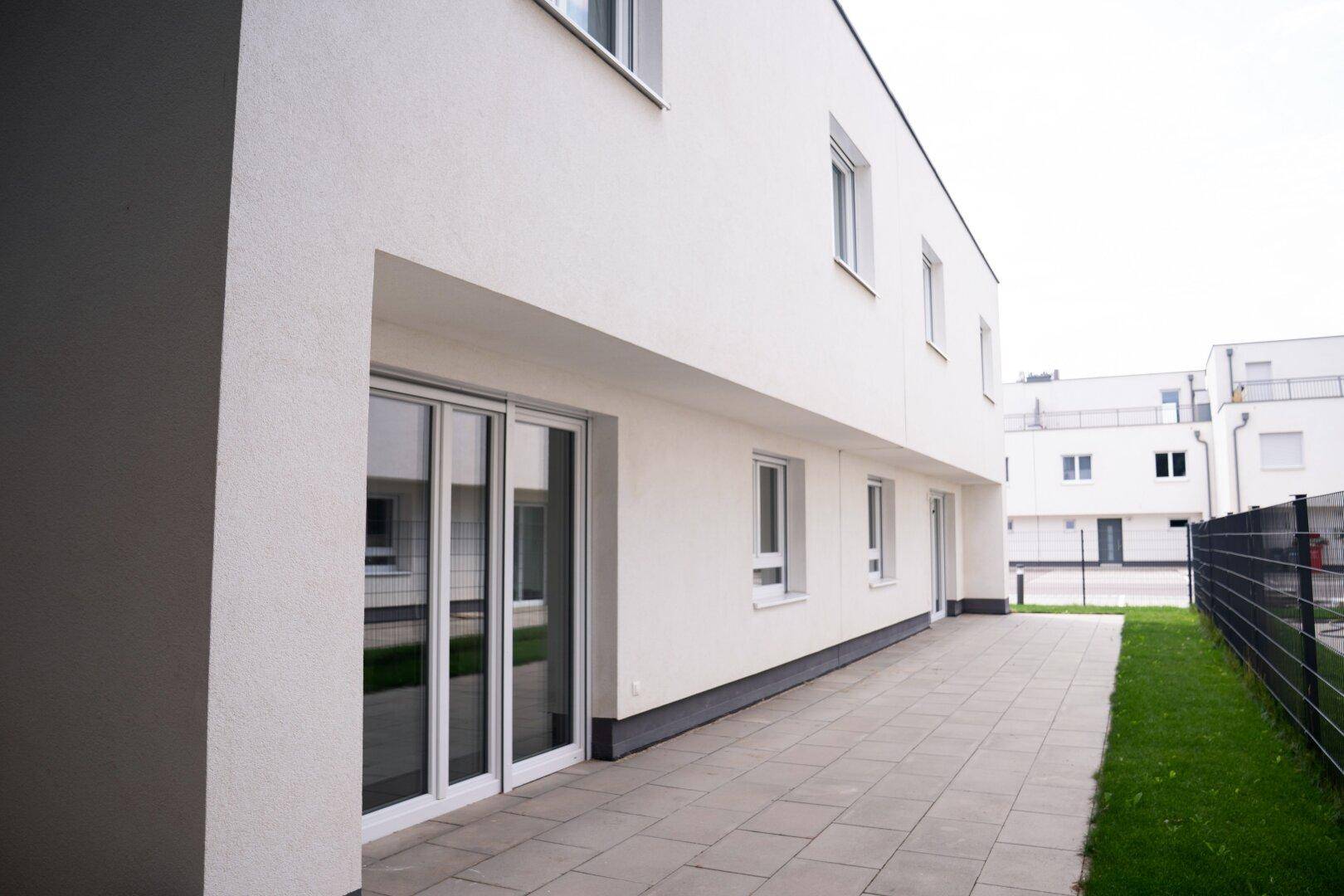 Seitliche Ansicht eines modernen Gebäudes mit gepflasterter Terrasse und großen Schiebetüren zum Garten.