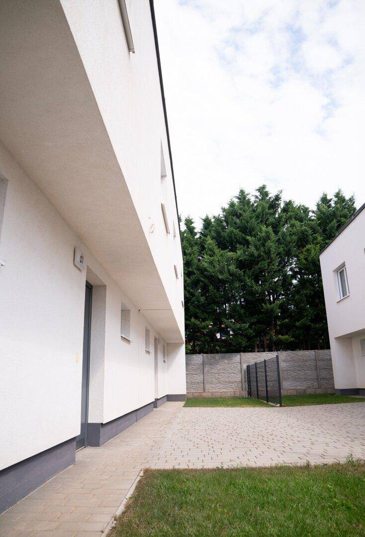 Moderne Gebäudefassade mit gepflastertem Weg, Rasenfläche und hohen Bäumen im Hintergrund der Anlage.