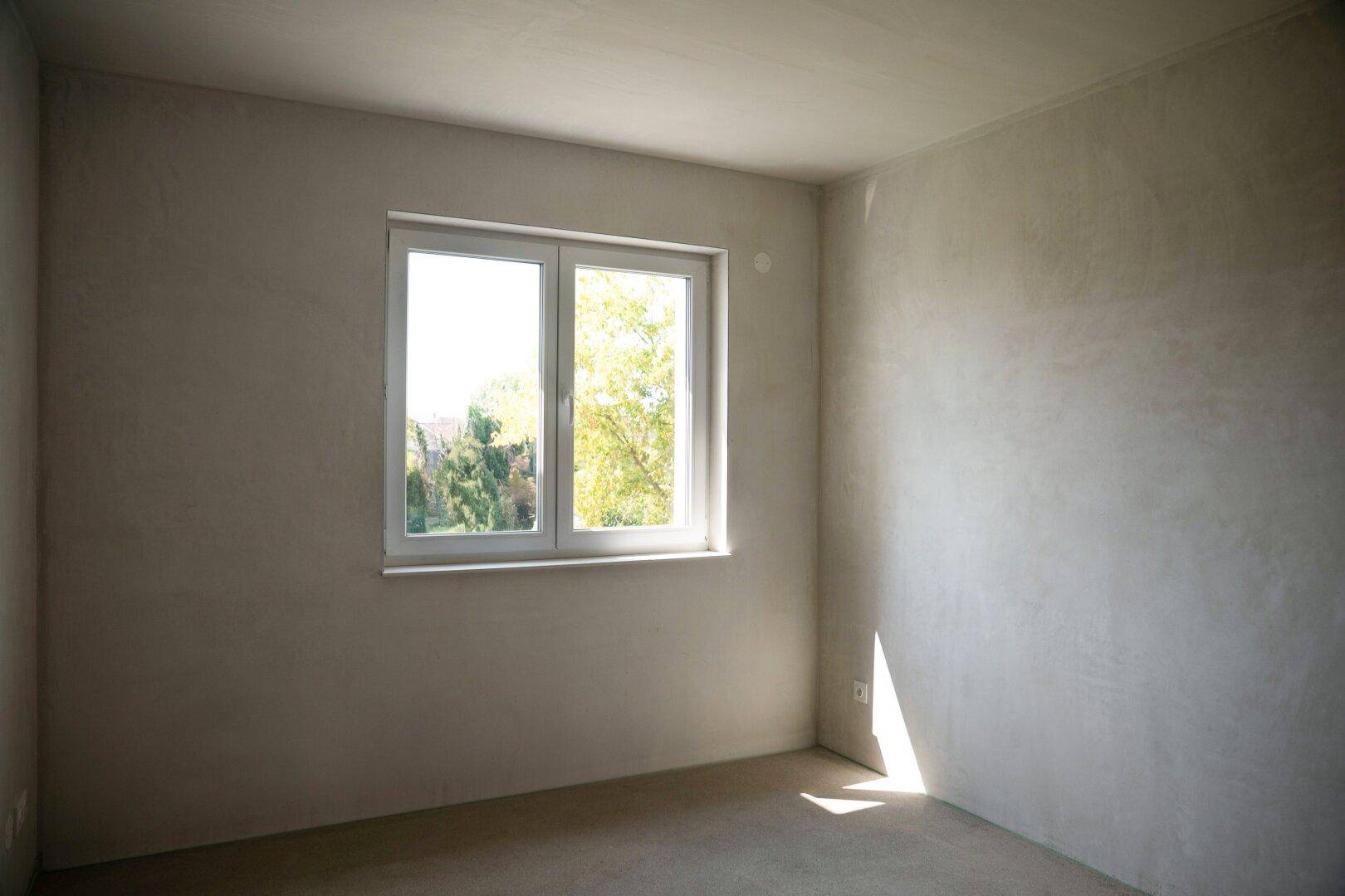 Heller Raum im Rohbau mit Fenster und Blick auf grüne Bäume, ideal als zukünftiges Schlafzimmer.