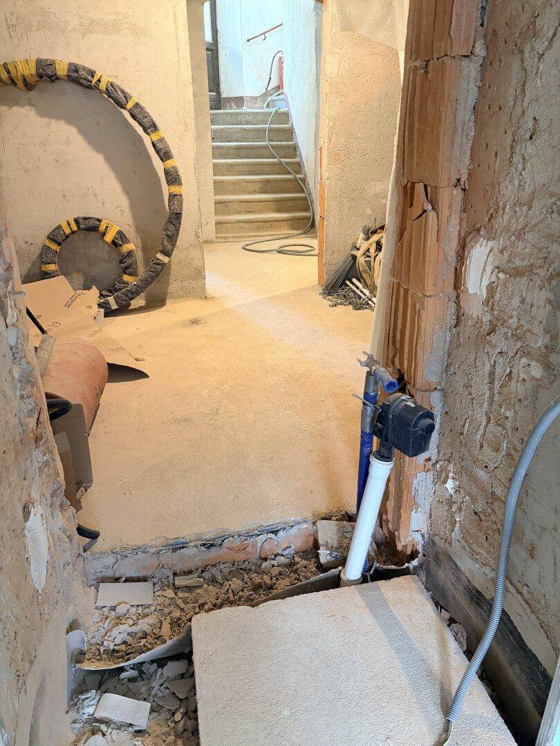 Kellerbereich im Rohbau mit sichtbaren Leitungen, Betonboden und einer Treppe im Hintergrund.