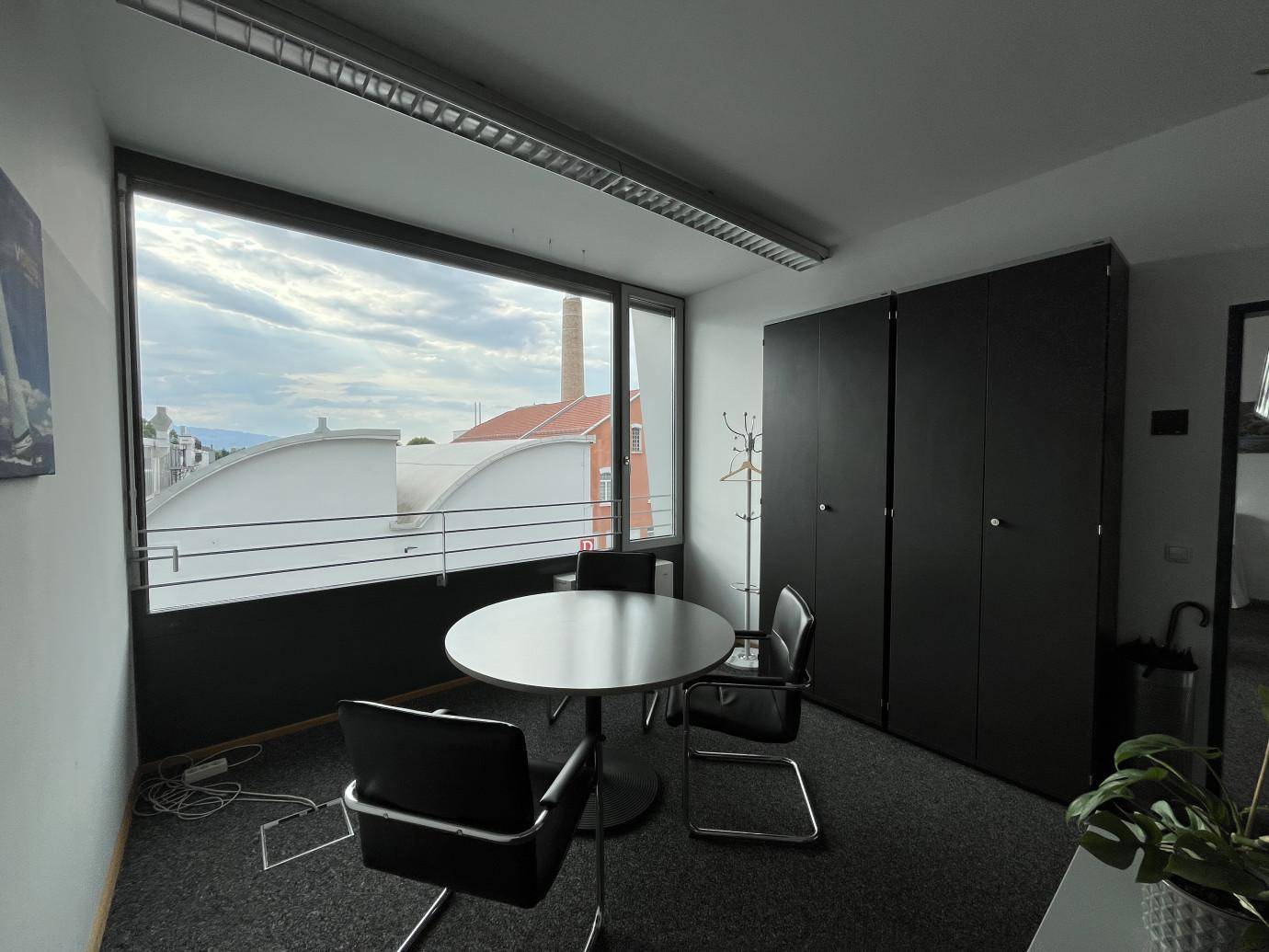Büro mit Besprechungstisch, dunklen Schränken und Blick auf Dächer und einen Schornstein.