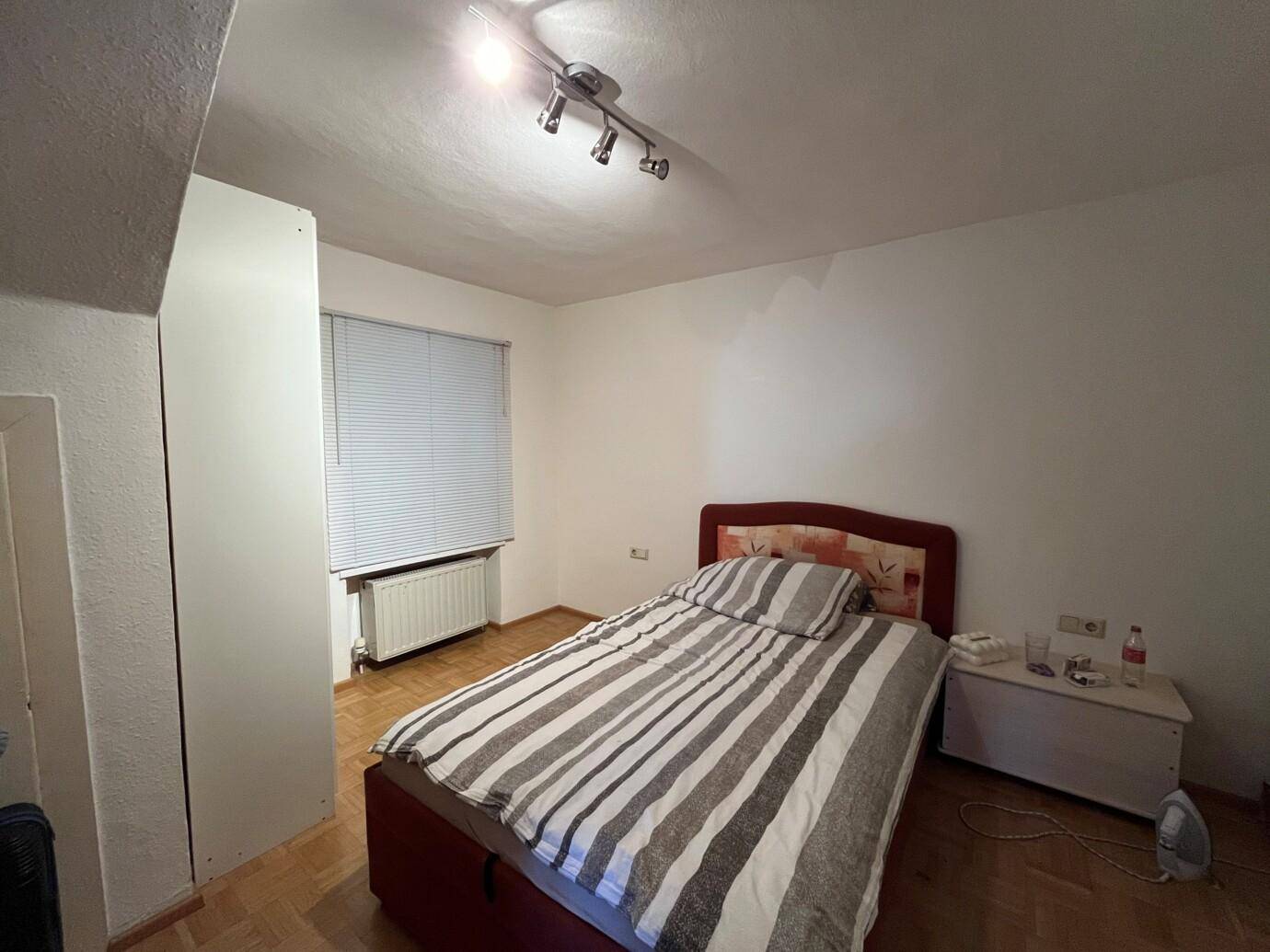 Ein bescheidenes Schlafzimmer mit Ehebett, brauner Bettwäsche und gestreifter Decke. Ein weißer Nachttisch steht daneben.
