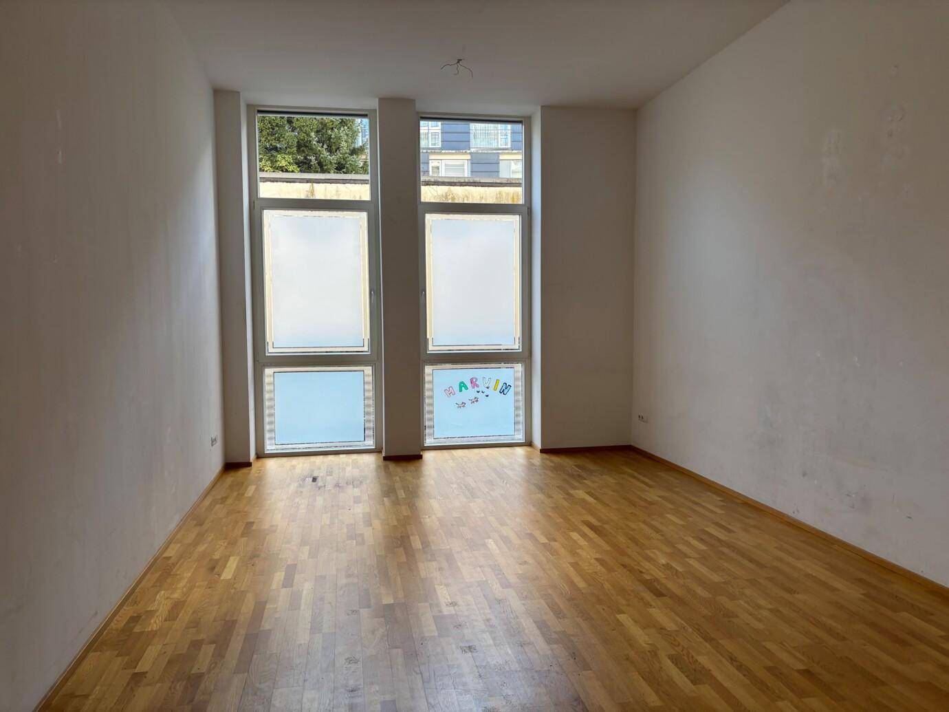 Heller, leerer Raum mit Holzparkett und großen Fenstern, die Tageslicht spenden.