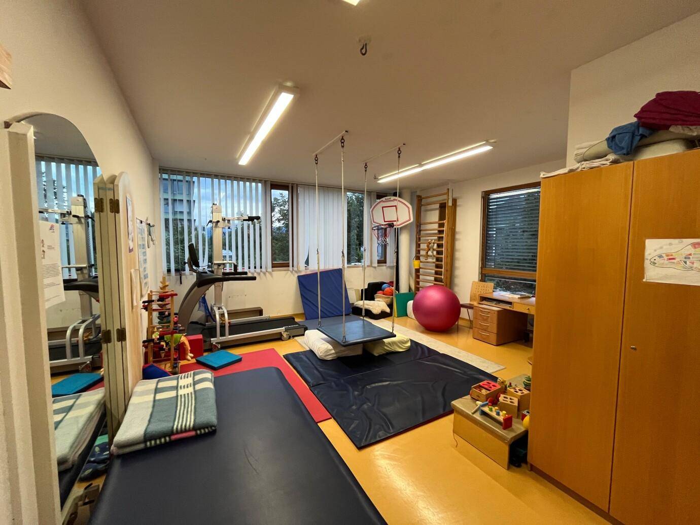 Großer Therapieraum mit Sportgeräten, Matten, Schaukel und Fenstern mit Blick auf die Umgebung.